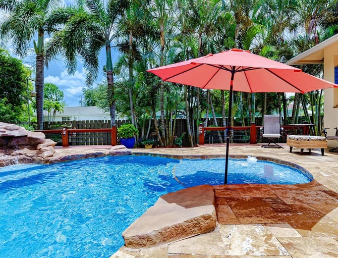 Backyard swimming pool with red umbrella, lounge chairs, and palm trees surrounding patio area